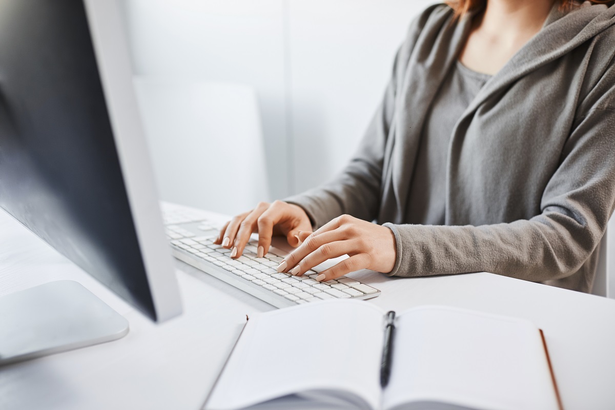 Working Hard To Party Hard On Weekends. Cropped Shot Of Woman Typing On Keyboard, Sitting In Front Of Computer Monitor. Freelancer Translating New Project, Writing Some Notes In Note Pad.